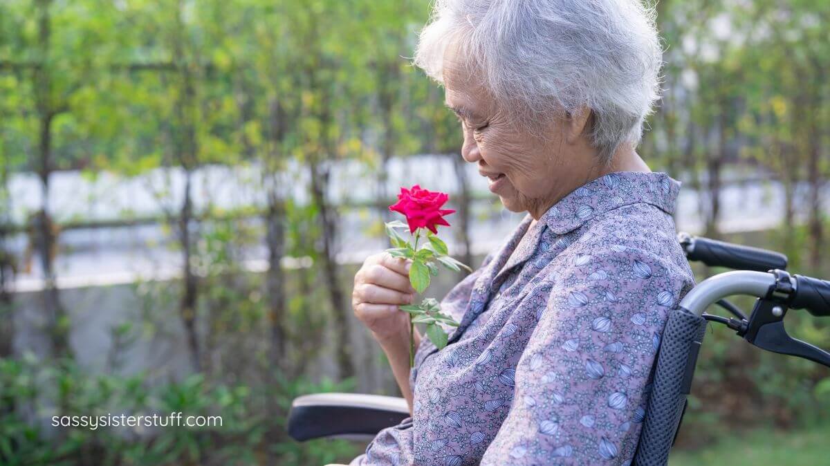 senior female sitting outside in her wheel chair in a rose garden holding a rose and smiling at it.