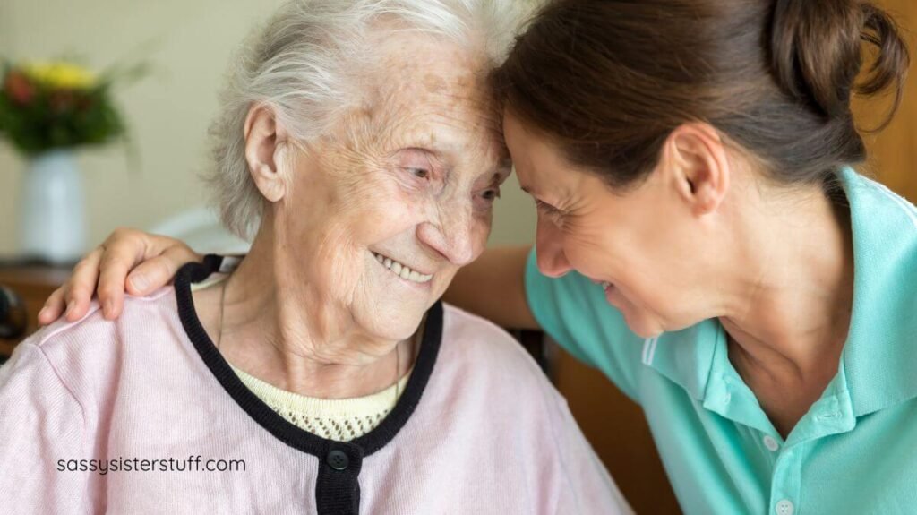 a caregiver and senior woman who has dementia sit head-to-head smiling at each other.