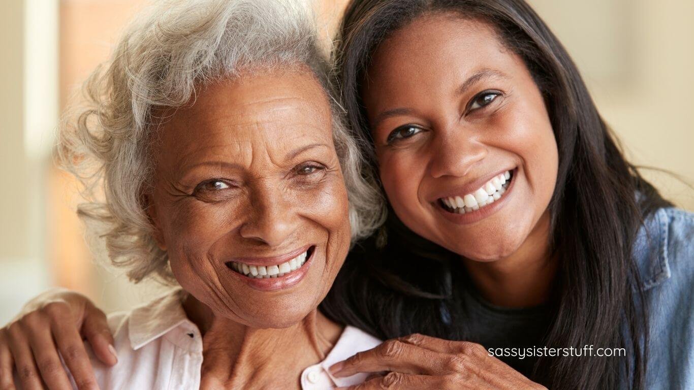 an elderly mother and her adult daughter with their heads together smiling at the camera.