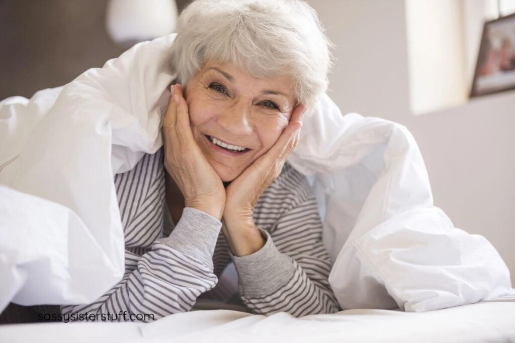 senior woman under the covers of her bed with her chin resting in her hands laughing at someone.