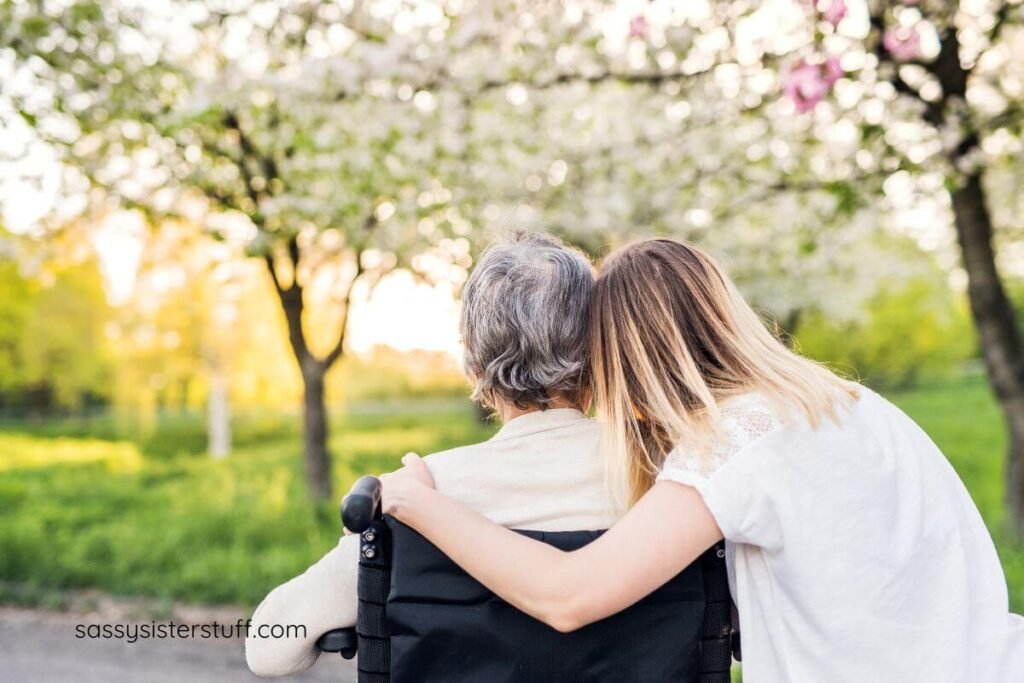 adult woman hugs her elderly mother who is sitting in a wheelchair looking across a park in the spring.