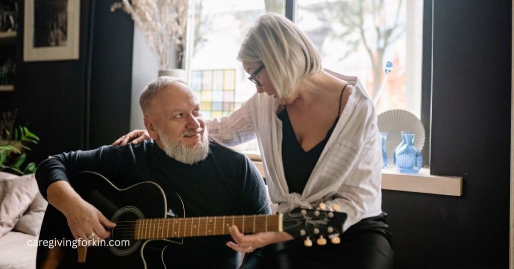wife has her arm around the shoulders of her husband who has dementia but is trying to play his guitar.