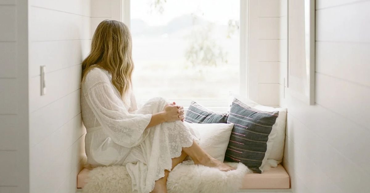 woman sits on a window seat looking out the window because she is tired of caregiving and dealing with anticipatory grief.